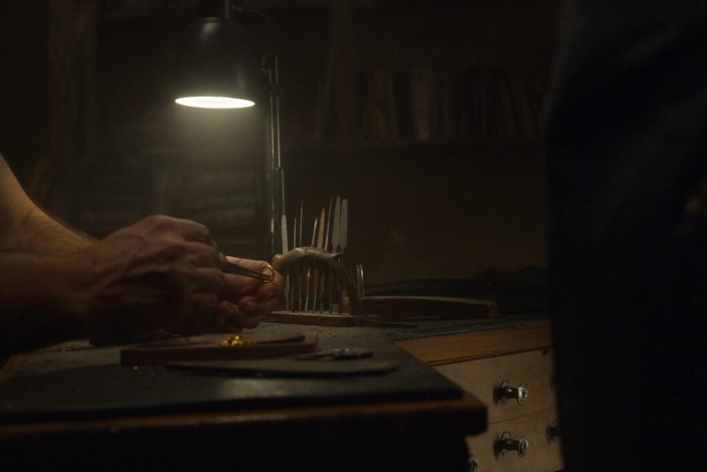 Hands holding a gold ring with tweezers at a jeweller's workbench under a warm lamp