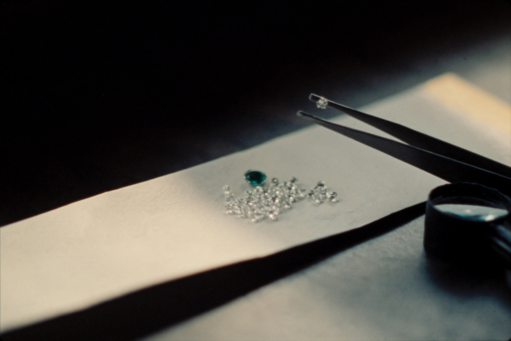 Loose diamonds and an emerald on a paper parcel with tweezers and a loupe