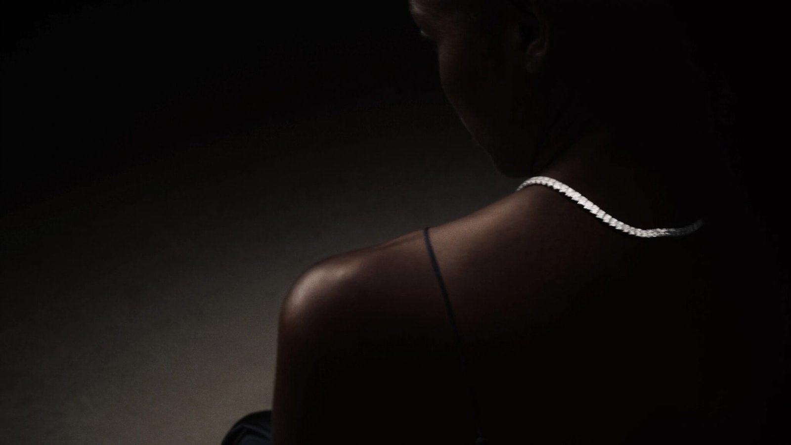 A woman in a dark silk dress seen from behind wearing a diamond riviera necklace in low light