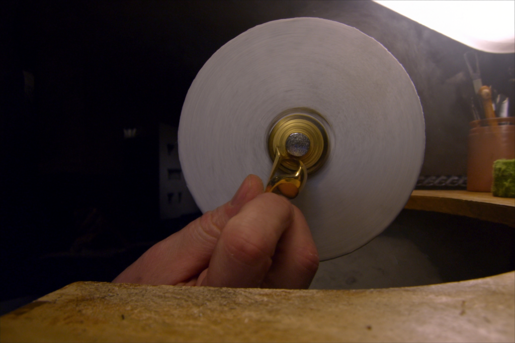 A hand polishing a gold ring against a rotating buffing wheel at the bench